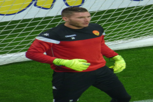 Ein Mann in rotem Hemd und schwarzen Hosen steht auf einem Fußballfeld mit einem Netz und einer Fahne im Hintergrund, trägt Fußballhandschuhe.