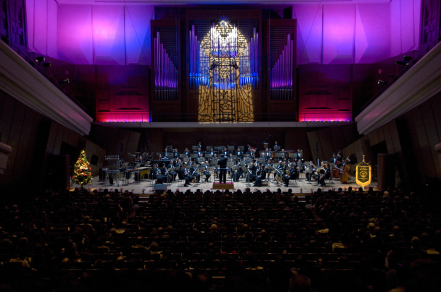 Ein voller Saal mit Menschen, die ein Konzert genießen, mit Musikern auf der Bühne und einem geschmückten Weihnachtsbaum im Hintergrund.