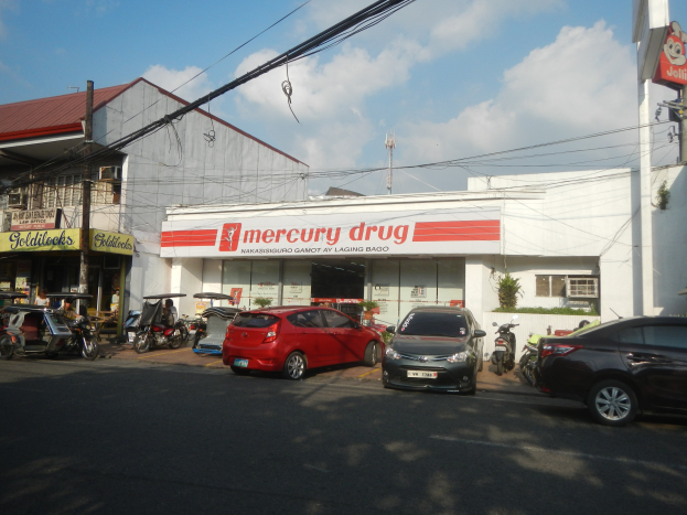 Eine belebte Straße mit Fahrzeugen, Gebäuden, Strommasten und Wolken im Hintergrund, mit einem Mercury Drug Store im Vordergrund und parkenden Autos.