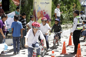 Kinder fahren Fahrräder auf einer Straße mit Verkehrskegeln, einige tragen Helme, andere stehen daneben; ein Banner mit Text, Bäume und Gebäude im Hintergrund.