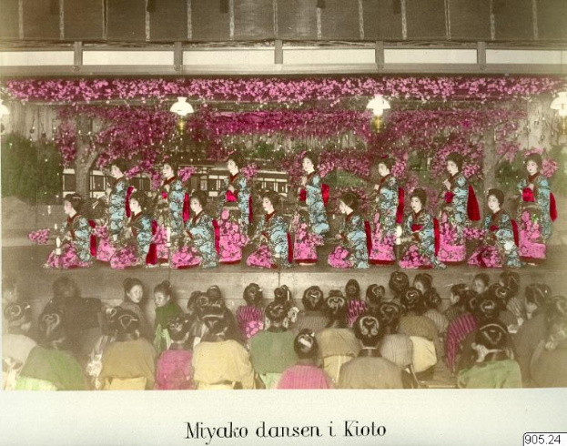 Ein altes Foto einer Gruppe von Frauen in leuchtenden Kimonos, die auf einer mit Blumen und Lichtern geschmückten Bühne sitzen, mit einem Publikum in Stühlen und Text am unteren Bildrand.