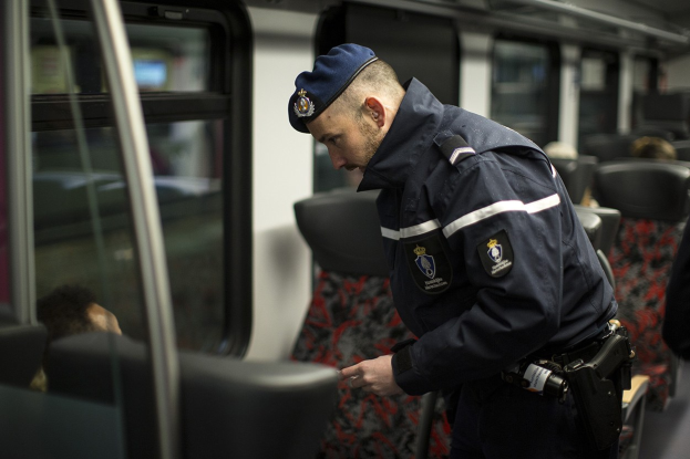 Polizist in Uniform und Mütze auf einem Zug stehend, der auf sein Handy schaut, mit sitzenden Passagieren und einer weiteren stehenden Person im Bild.