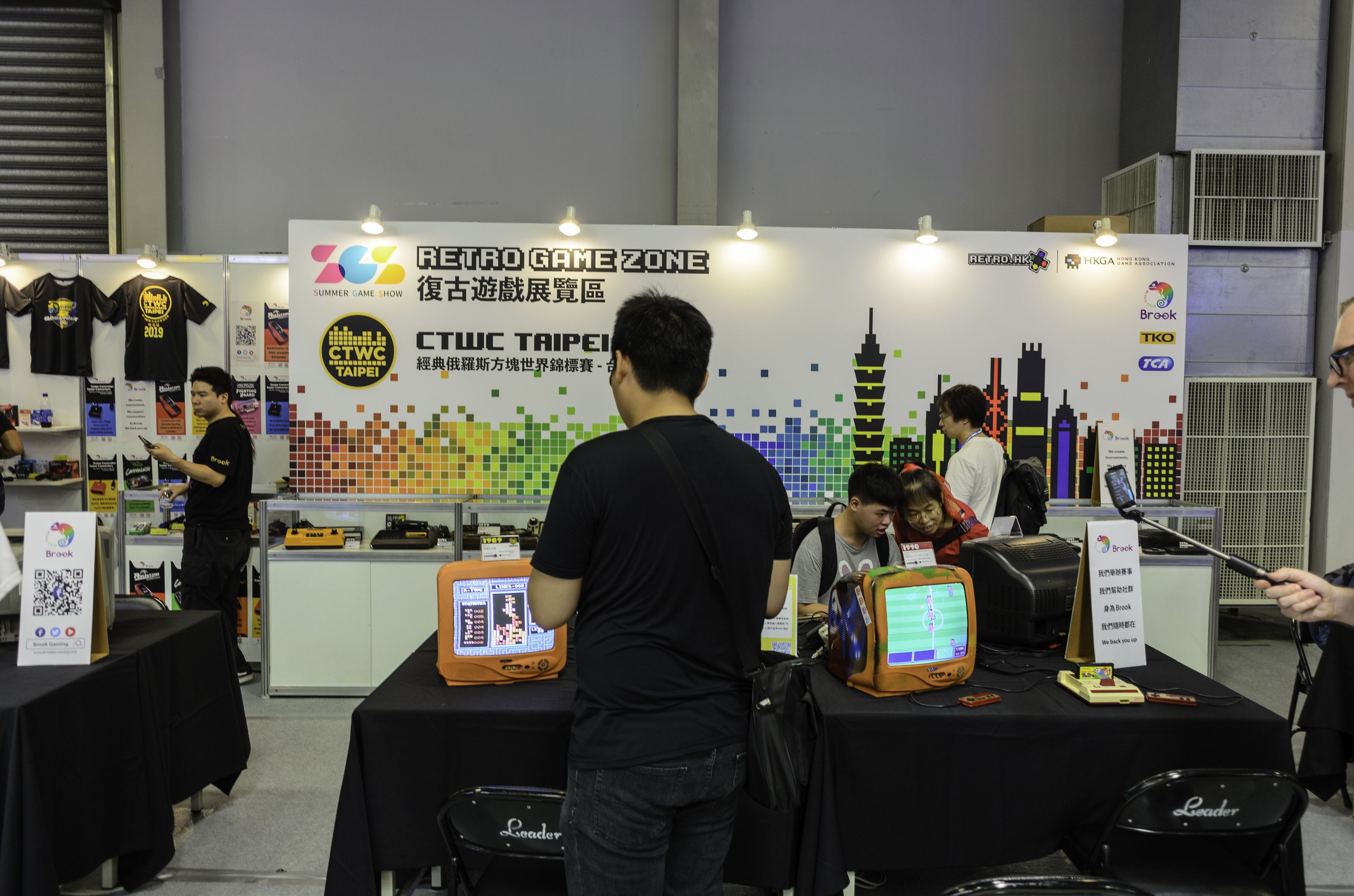 Leute sitzen um einen Tisch und spielen ein Videospiel, Stühle, eine Wand, aufgehängte T-Shirts und Deckenbeleuchtung sind zu sehen.