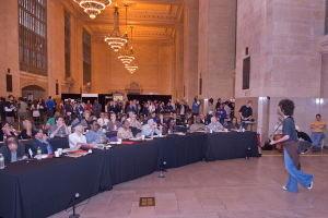 Eine Konzertszene mit einer Menge Menschen, einem Mann, der rechts Gitarre spielt und singt, einer Wand und Fenstern im Hintergrund, Lampen oben und einem Boden unten.