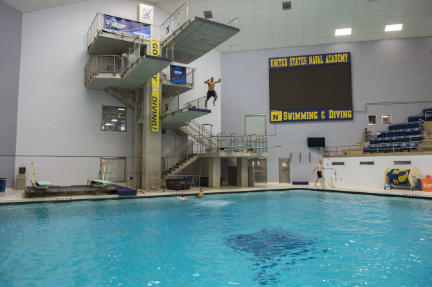 Großer Hallenbad mit schwimmenden Menschen, Treppe mit Geländer, Banner mit Text, Person, die ins Wasser springt, Stühle rechts, Bildschirm an der Wand und Deckenbeleuchtung.