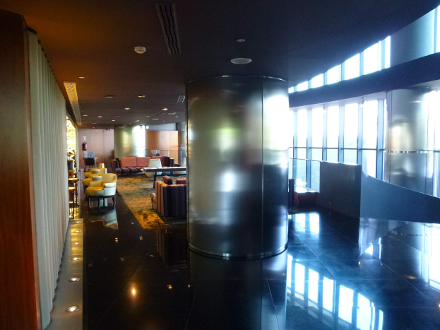 Eine geräumige Business-Lounge am Hong Kong International Airport mit großen Fenstern, hohen Decken mit elektrischem Licht, Sitzmöglichkeiten einschließlich Stühle und Sofas, Tischen, einem Teppichboden und einer zentralen Säule.