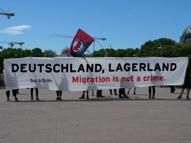 Eine Gruppe von Menschen mit einer Fahne und einem Banner mit der Aufschrift "Deutschland, Lagerland Migration ist kein Verbrechen" sowie Straßenlaternen, Bäumen, Kränen, einem Gebäude und einem bewölkten Himmel im Hintergrund.