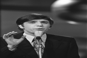 Ein Mann in einem schwarzen Anzug, weißem Hemd und gemustertem Schlips spricht ernst in ein Mikrofon und hat sein Haar zurückgek├Ąmmt.