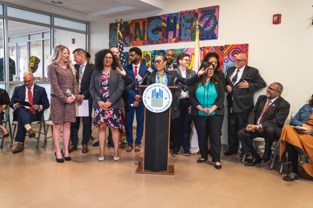 Eine Gruppe von Menschen, die vor einem Podium mit einem Mikrofon und einer Tafel mit der Aufschrift "Sancho-Stadtrat tritt dem Stadtrat bei" stehen und sitzen. Im Hintergrund sind Flaggen, ein Feuerlöscher und Deckenleuchten zu sehen.