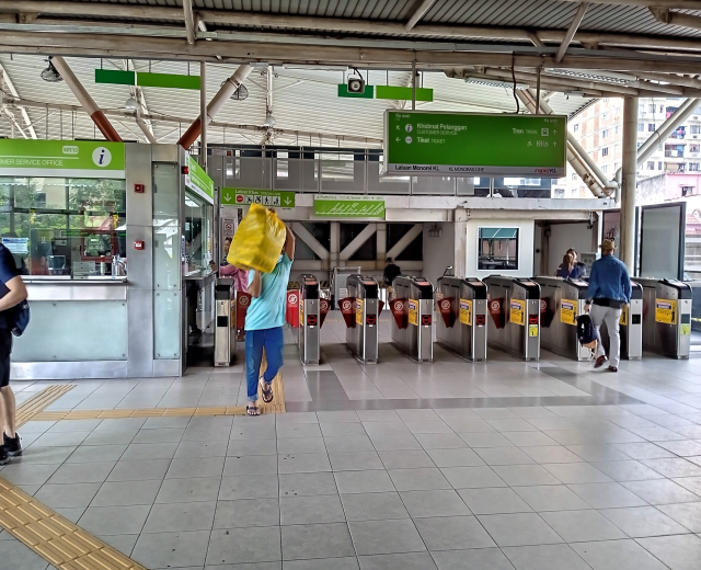 Menschen gehen durch einen Bahnhof mit Metalldetektoren, Hinweisschildern, Metallstangen, Deckenlampen und Gebäuden mit Fenstern im Hintergrund.