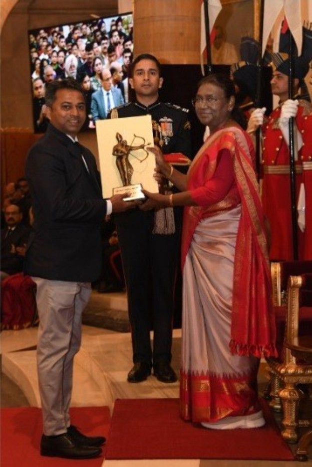 Ein Mann in einem schwarzen Blazer, weißem Hemd und grauen Hosen, der eine Trophäe hält, neben einer Frau in einem aschfarbenen Sari auf einem roten Teppich bei einer Preisverleihung, mit Menschen, Stühlen, Flaggen und einem Bildschirm im Hintergrund.
