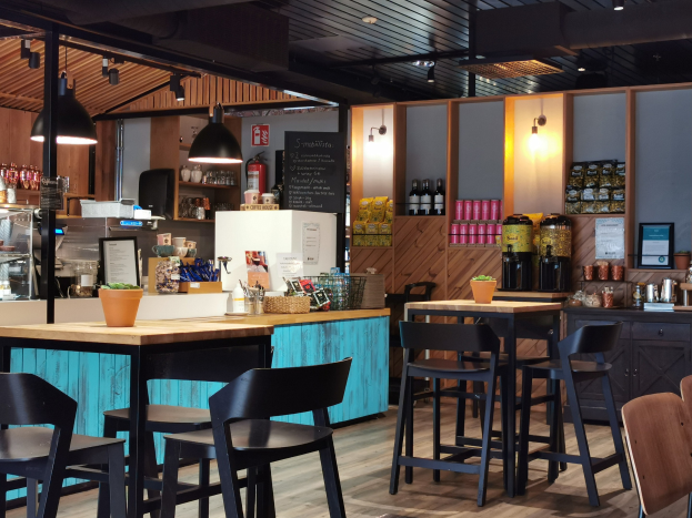 Innenraum eines Restaurants mit Holztischen, -stühlen, einer Topfpflanze auf einem Tisch, einer Schiefertafel, einem Kühlschrank, einem Feuerlöscher, Regalen mit Flaschen, Schränken mit Gegenständen, einem Rahmenfoto an der Wand und Deckenlampen.