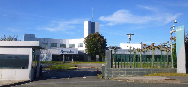Ein großes Gebäude mit zahlreichen Fenstern und einem Tor davor, umgeben von Straßenmasten, Laternen, Gittern, Bäumen und Gras, mit einem bewölkten Himmel im Hintergrund, das als Hauptsitz von Iberostica in den Niederlanden dient.