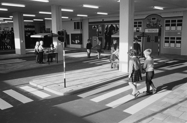 Schwarz-weißes Foto von Kindern, die eine Straße an einer Ampel überqueren, mit Menschen auf Bänken, einem Verkehrssignalmast im Vordergrund und Säulen mit Lampen im Hintergrund.