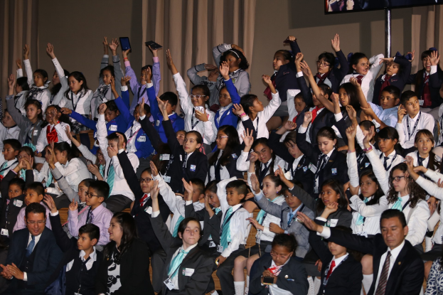 Gruppe von Kindern sitzt auf einer Bühne mit einem Mann und einer Frau davor; Bühnenhintergrund enthält Vorhänge, eine Wand und einen Fernseher auf einem Ständer.