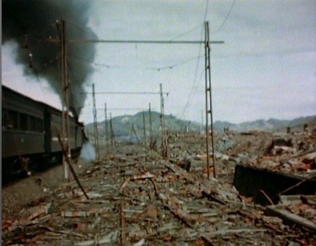 Ein Zug fährt durch eine verwöstete Landschaft mit Rauch, der aus ihm aufsteigt, umgeben von Strommästen und -drähten, mit Höhen und einem klaren Himmel im Hintergrund und verstreuter Trömmerei am Boden.