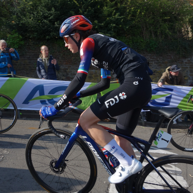 Eine professionelle Radrennfahrerin, Katie Archibald, trägt einen Helm, eine Brille, Handschuhe und Schuhe und fährt ein Fahrrad eine Straße entlang neben einer Menge, mit einem Banner, einer Wand, Bäumen und einem Gebäude im Hintergrund.