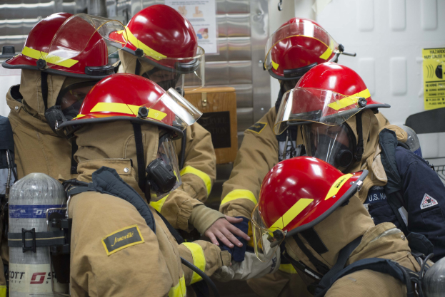 Feuerwehrleute in Schutzausrüstung mit Sauerstoffflaschen arbeiten zusammen, um ein Feuer zu löschen, mit Plakaten im Hintergrund.