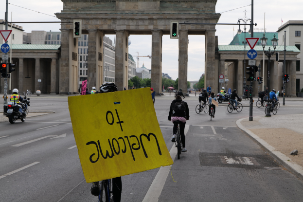 Eine Gruppe von Menschen fährt Fahrräder vorbei am Brandenburger Tor in Berlin, Deutschland, mit einer Person, die ein gelbes Schild hält, unter einem klaren Himmel mit Bäumen und Gebäuden im Hintergrund.
