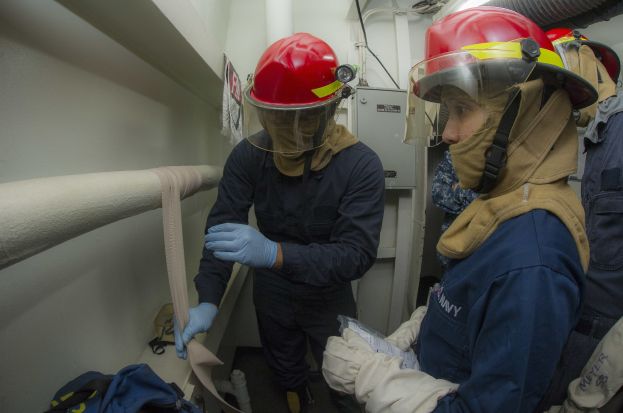 Drei Personen in Feuerbekämpfungsausrüstung stehen zusammen in einem Raum mit Rohren, einer Tasche und Maschinen im Hintergrund.