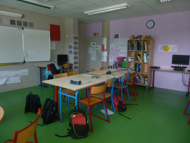 Ein Klassenzimmer mit Tischen, Stühlen, Taschen, Bücherregalen, einem Monitor, Tastatur, Maus, Uhr, Papieren an den Wänden, einer Tafel und Deckenleuchten.