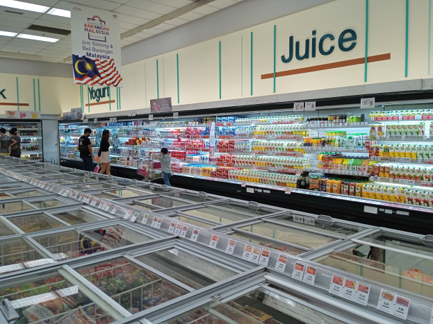 Ein gut sortierter Lebensmittelgang mit Regalen voller Getränke, Gemüse und anderen Lebensmitteln, ein paar Einkäufern und Deckenbeleuchtung.