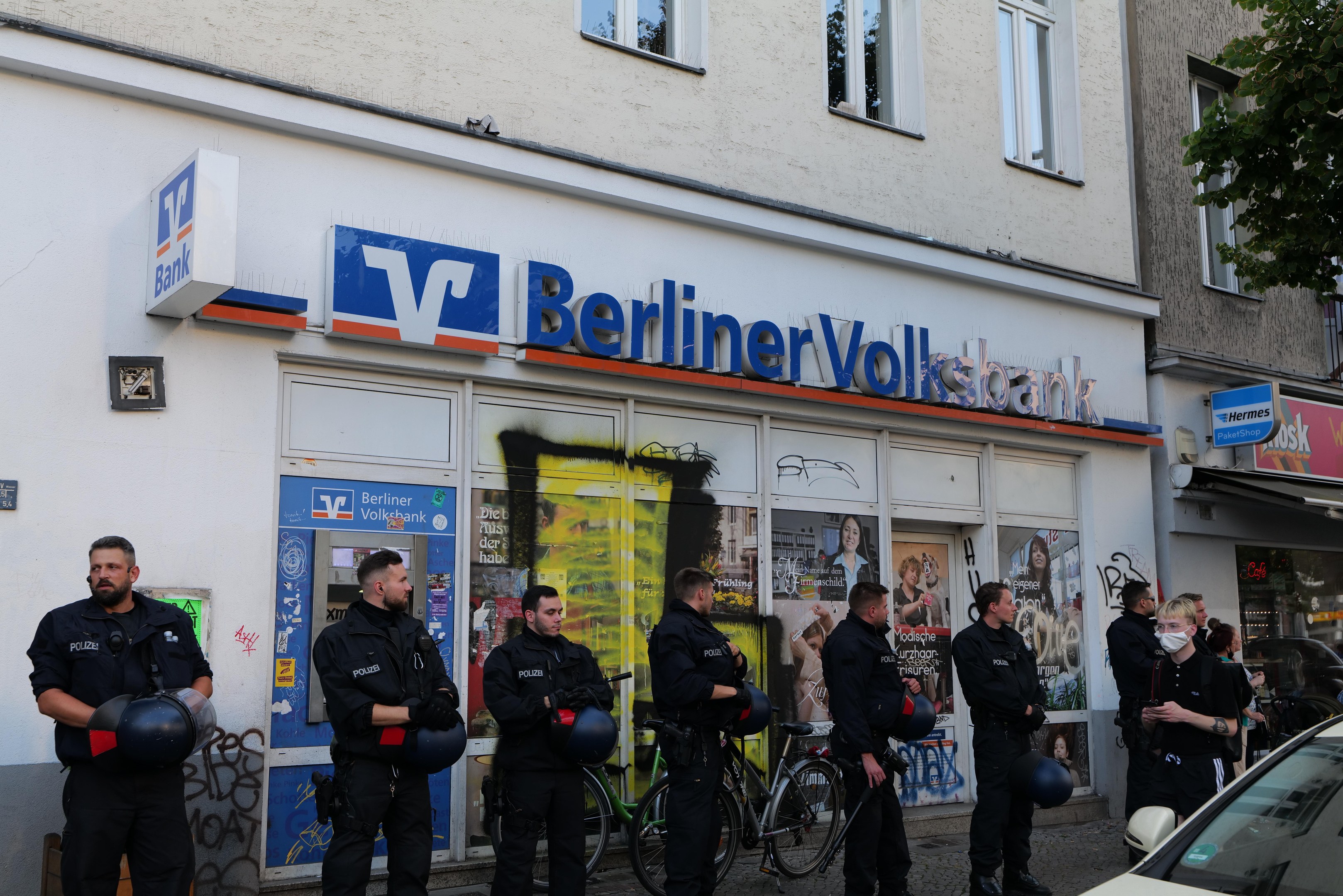 Polizeibeamte in Uniform stehen vor einer Berliner Volksbank-Filiale, einige halten Helme, mit einem geparkten Auto rechts und einem Baum links, vor einem Hintergrund von Gebäuden mit Fenstern und textbedeckten Schildern.