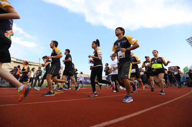 Eine Gruppe von Menschen, die auf einer Laufbahn laufen, mit einer Zuschauermenge auf der rechten Seite, vor Gebäuden, Laternenmasten, Bannern und einem klaren blauen Himmel während eines Rennens.