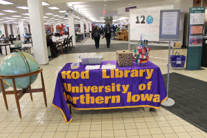 Eine Ausstellung mit einem Tisch, auf dem verschiedene Landesflaggen, eine Box, Papiere und ein Banner sowie links daneben eine Weltkugel, rechts eine Anschlagtafel mit ein paar Menschen in der Nähe und mehrere Tische und Stühle zu sehen sind.