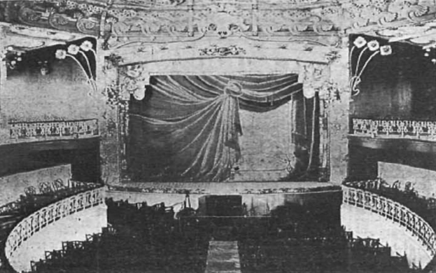 Schwarzes und weißes Foto des Auditoriums des Theatre Royal in London, das eine Bühne mit einem Vorhang, Geländern und ornamentierten Wanddesigns zeigt, sowie ein paar sitzende Menschen und Text am unteren Rand.