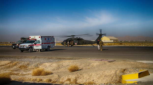 Helikopter neben einem Rettungswagen auf einer grasbewachsenen Straße geparkt mit Menschen in der Nähe, Hügel und einen klaren blauen Himmel im Hintergrund.