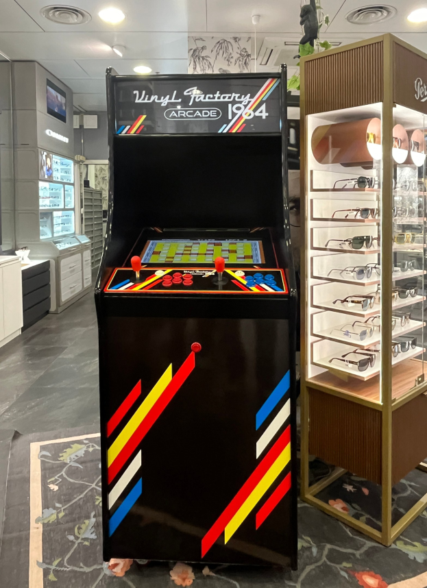 Ein Geschäft mit einem zentralen Retro-Arcade-Automaten, einem rechten Gestell mit Gläsern und Hintergrundschränken, Fernseher und anderen Gegenständen sowie Deckenleuchten, die eine Retro-Atmosphäre schaffen.
