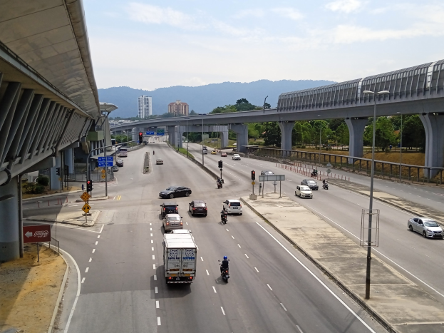 Eine vielbefahrene Autobahn mit mehreren Fahrzeugen, neben einer Brücke, mit Straßenlaternen, Verkehrszeichen, Schildern, Bäumen, Gebäuden, Hügeln und einem bewölkten Himmel.