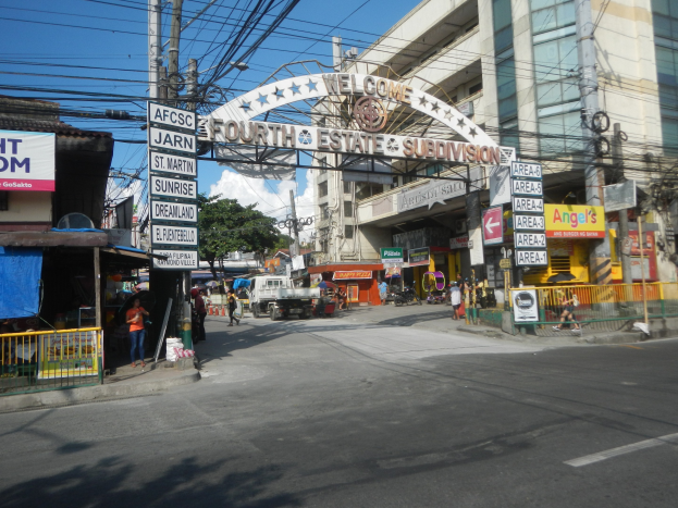 Eine belebte Straße mit fahrenden Fahrzeugen, Füßgängern auf dem Gehweg, einem Tor mit Text, Gebäuden mit Fenstern, Strommästen mit Überlandleitungen, Bäumen und einem bewölkten Himmel, mittig von einem Straßenschild mit der Aufschrift "Fourth Estate Subdivision".