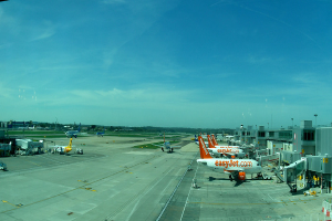 Eine Flughafen-Szene mit Flugzeugen, Gebäuden, Polen, Fahrzeugen, Bäumen, Himmel, Straße und einem Zaun im Vordergrund.