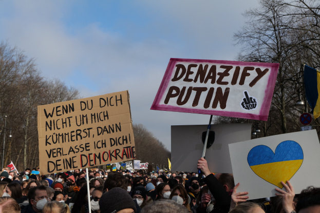 Eine große Gruppe von Menschen hält Schilder und Fahnen mit der Aufschrift "Denazifizieren Sie Putin" während einer Demonstration in Deutschland, mit Bäumen und einem klaren Himmel im Hintergrund.