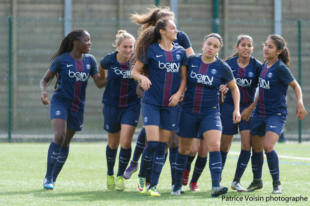 Eine Gruppe junger Frauen, die auf einem Rasenfeld Fußball spielen, mit einem Maschendrahtzaun und Pfosten im Hintergrund und einer Wand dahinter, mit dem Text "Paris Saint-Germain Frauenfußball" in der rechten unteren Ecke.
