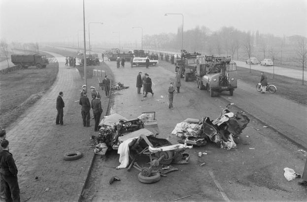 Schwarz-weißes Bild einer Autounfall-Szene am Straßenrand mit mehreren Fahrzeugen, einer Gruppe von Menschen in der Nähe und Laternen, Bäumen und Himmel im Hintergrund.