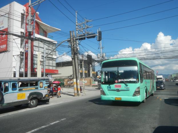 Ein grüner Bus fährt auf einer Straße mit hohen Gebäuden entlang, mit Fußgängern, die Regenschirme halten, und Strommasten mit Drähten an der Straße unter einem bewölkten Himmel.