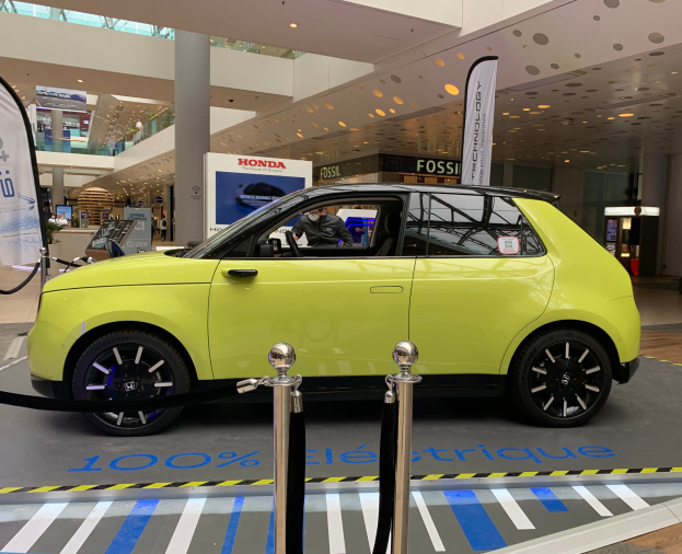 Ein gelbes wasserstoffbetriebenes elektrisches Auto vor einem Gebäude geparkt, umgeben von Absperrpollern mit Seilen und Schildern, mit einer Person im Auto, einer Säule, einer Gruppe von Menschen, Schildern, einem Topf mit einer Pflanze, Deckenleuchten und einer Decke mit Deckenventilatoren.