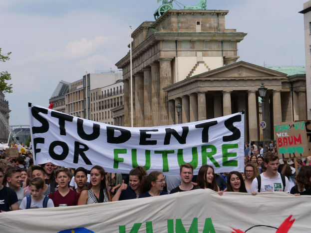 Gruppe von Schülern marschiert in Berlin mit einem leuchtend bunten "Students for Future"-Schild vor einer Kulisse aus Gebäuden, Bäumen und Himmel.