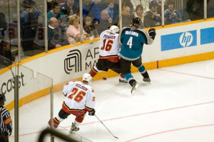 Ein Mann in einem sechzehn Nummern-Jersey, Helm, Shorts und Schuhen steht neben einem anderen Mann in einer blauen Jacke, Brille, Helm und Hose neben einem Fenster, mit einem Publikum darüber, das ein Spiel beobachtet, einem Mann in einem sechsundzwanzig Nummern-Jersey, der einen Stock hält, einem Schiedsrichter hinter Glas in der unteren linken Ecke und einer Werbetafel rechts.