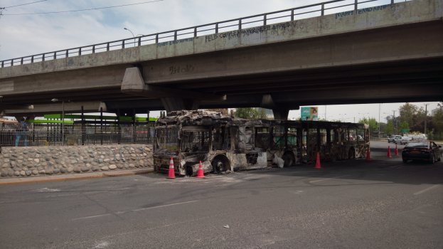 Vernichteter Bus am Straßenrand mit Absperrkegeln, umgeben von einem Zaun, Bäumen, Straßenmasten, einem Schild, einer Brücke, Drähten und einem bewölktem Himmel.
