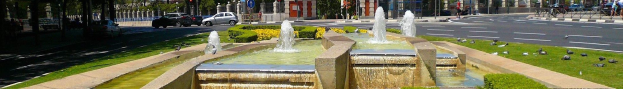 Ein Brunnen auf einer Stadtstraße umgeben von Grün, mit Fahrzeugen und Fußgängern, vor einem Gebäude- und klaren Himmel.