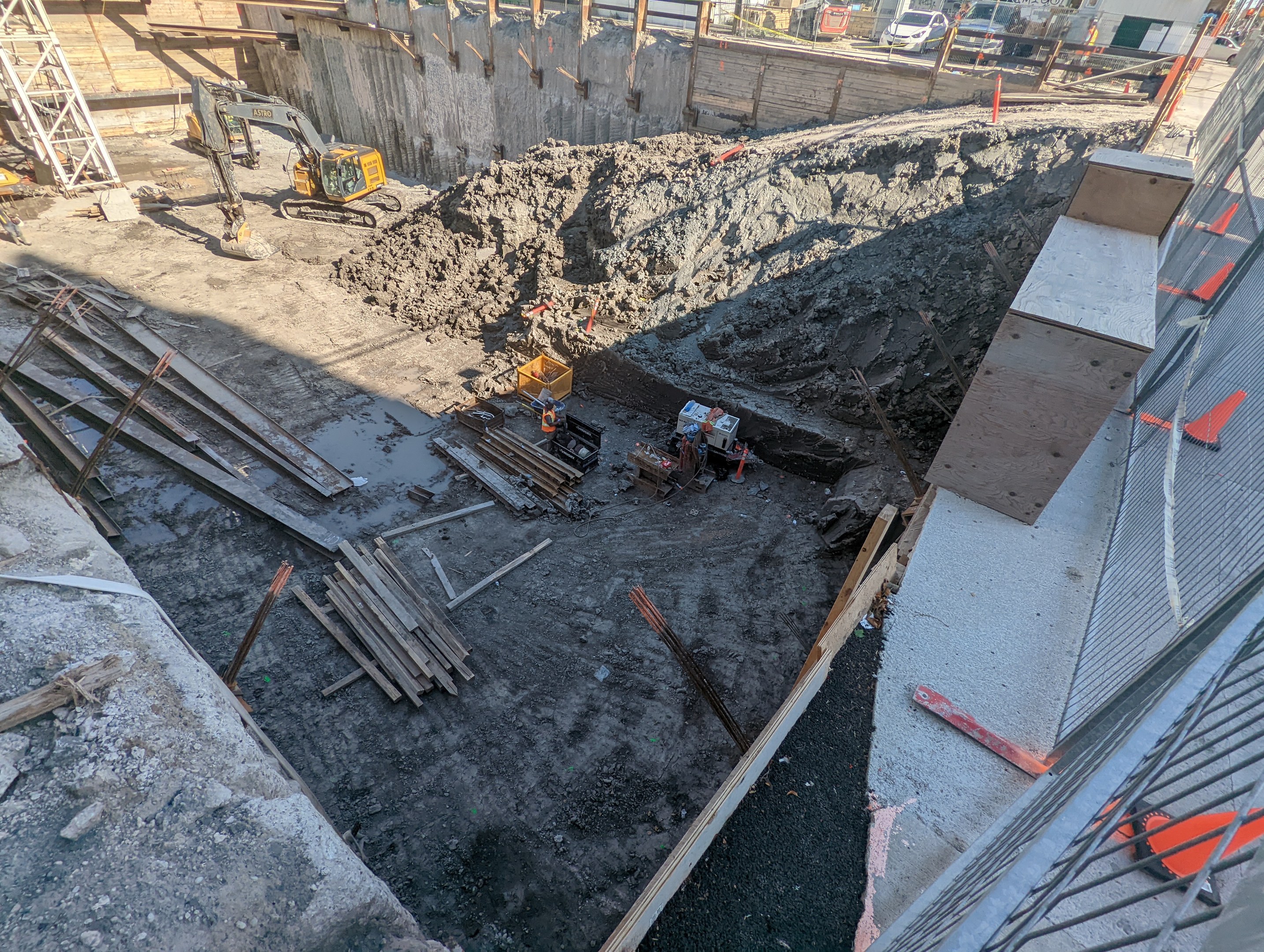 Baustelle mit einem großen Haufen Erde, umgeben von Kränen, Holzstöcken und anderem Equipment, begrenzt von einem Maschendrahtzaun auf der rechten Seite, mit Gebäuden, Fahrzeugen, Pfählen, Bäumen und Himmel im Hintergrund.