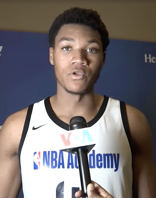 Ein Mann in einem weißen Tanktop hält ein Mikrofon und spricht vor einem Banner, auf dem "NBA All-Star Game 2019" steht und einer Wand im Hintergrund.