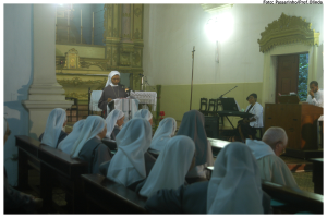 Eine Frau steht auf einem Podium, während Menschen auf Bänken und Stühlen im Hintergrund sitzen.