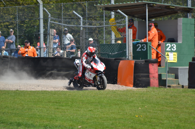 Eine Person in Helm und Handschuhen fährt ein Motorrad über ein grasbewachsenes Feld, mit Zuschauern in Mützen und Schutzbrillen, einem eingezäunten Bereich, Treppen mit Geländern und einem nummerierten Schuppen im Hintergrund.