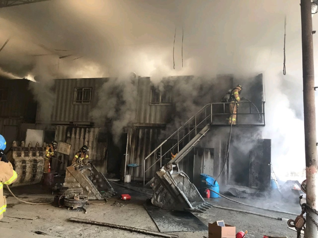 Feuerwehrleute in Helmen und Schutzausrüstung sprühen Wasser, um ein Lagerfeuer zu löschen, mit Rauch, der aus dem Gebäude aufsteigt und Trümmern, die auf dem Boden verstreut sind.