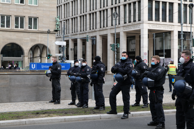 Polizisten in schwarzen Uniformen und Masken vor einem Berliner Gebäude mit Glasfenstern und Säulen, einige halten Helme, mit Laternenmästen, Ampeln, Schildern, einer Statue und Gras im Hintergrund.
